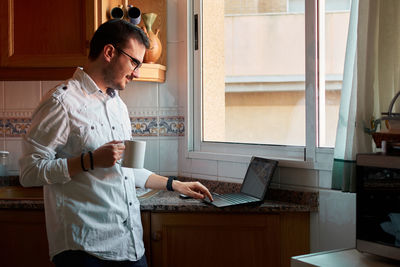 Young man looks at his laptop while drinking coffee in his kitchen