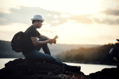 Man sitting on rock against sky during sunset