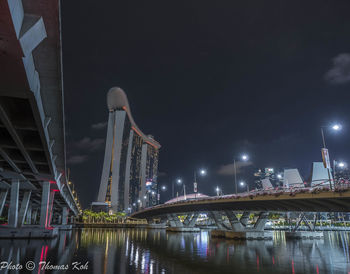 Illuminated bridge over river by buildings against sky at night