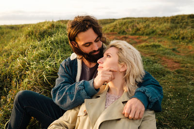 Bearded man touching face and embracing blond girlfriend while sitting on grassy hill on cloudy day in countryside in aviles, spain