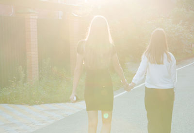 Rear view of friends holding hands while walking on road during sunny day