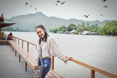 Portrait of smiling young woman standing by railing against sky