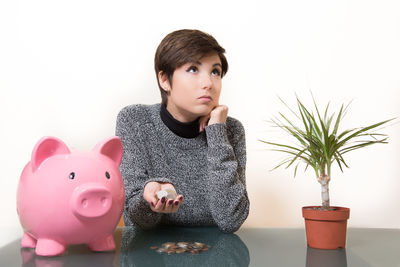 Young woman looking up while holding coins by piggy bank against wall