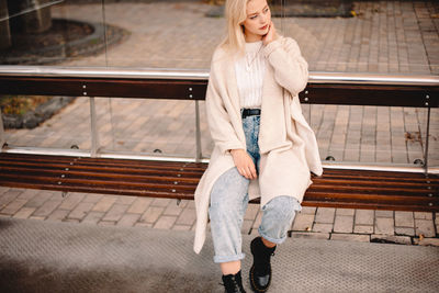 Thoughtful stylish young woman sitting on bus stop in city