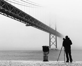 Full length rear view of photographer standing by 25 de abril bridge over tejo river during winter
