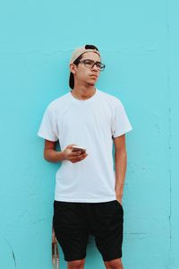 Thoughtful young man holding mobile phone standing against blue wall