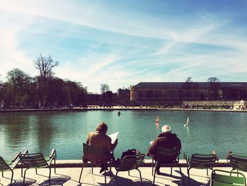Rear view of men sitting on chairs by lake against sky during sunny day