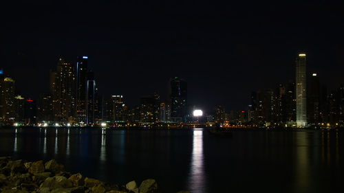 Illuminated buildings by river against sky at night