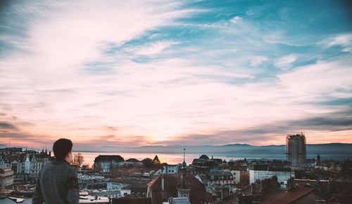 Rear view of man looking at city by lake against sky during sunset