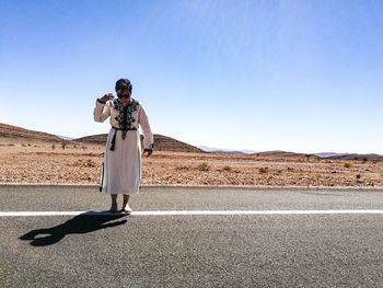 Woman standing on road at desert against clear blue sky