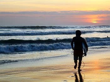 Rear view of man with surfboard running at beach during sunset