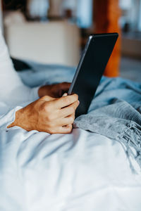 Side view of cropped unrecognizable guy touching screen of tablet while lying on bed in light modern apartment