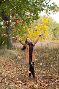 Woman throwing leaves while standing against trees in park during autumn