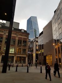 People walking on street by modern buildings in city against sky