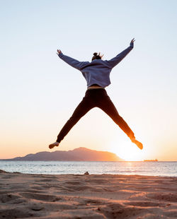 Rear view of woman jumping on beach against sunrise sky