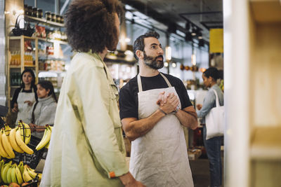 Mature sales clerk with hands clasped standing by male customer at grocery store