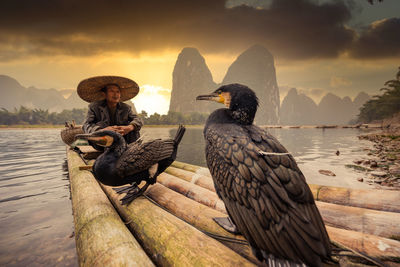 Man wearing hat sitting on raft with bird against mountains and sky