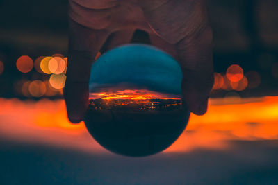 Close-up of man holding illuminated light bulb against sky at sunset