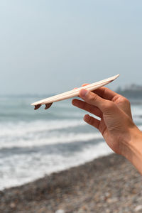 Cropped hand of person holding small surfboard against sky