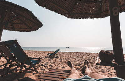 Low section of man relaxing on wooden deck chair at beach against sky