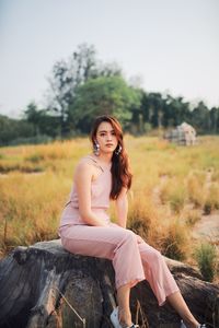 Portrait of young woman sitting on tree stump against clear sky