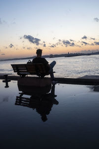 Rear view of man sitting on sea against sky during sunset