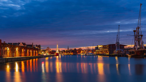 Illuminated buildings by river against sky at sunset