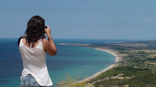 Rear view of woman photographing coastline against sky