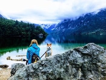 Rear view of mid adult woman with dog sitting on rock at lakeshore against cloudy sky