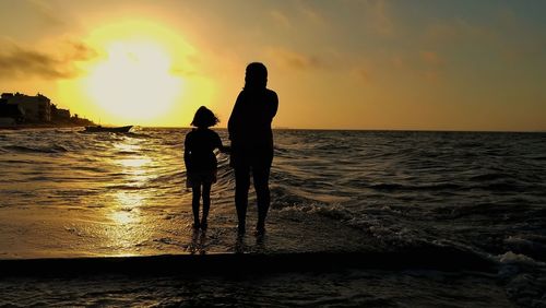 Silhouette woman with girl standing at beach against sky during sunset