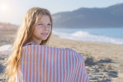 A young girl, covered with a towel, sits on a sandy beach. side view.