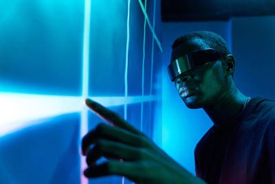 Concentrated young african american male in contemporary vr glasses touching large projector screen with geometric pattern while exploring cyberspace in dark room with neon illumination