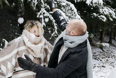 Mature couple with warm clothing having fun in snow by pine trees during vacations