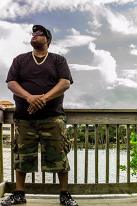 Mid adult man wearing sunglasses while standing by railing over river against cloudy sky