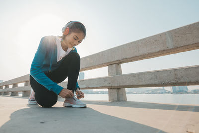 Full length of young woman tying shoelace by railing against sky