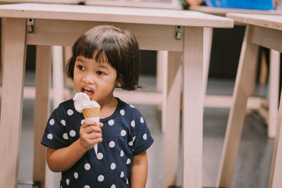 Cute girl eating ice cream while standing against tables