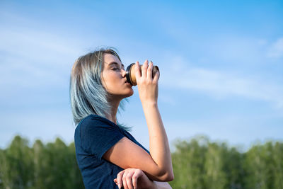 Blue haired woman drinks take away coffee outdoors on nature background.