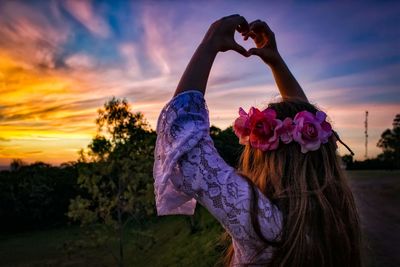 Woman wearing wreath making heart shape against cloud sky during sunset