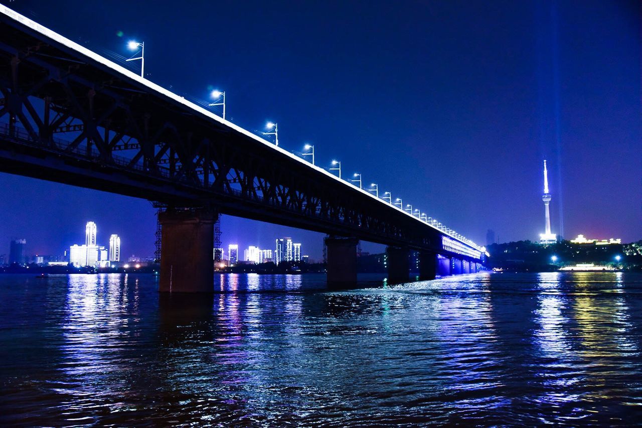 architecture, built structure, illuminated, connection, water, bridge - man made structure, night, waterfront, engineering, river, bridge, transportation, sky, city, suspension bridge, building exterior, reflection, blue, travel destinations, dusk