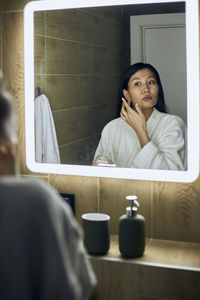 Woman wearing bathrobe applying moisturizing cream on face looking in mirror