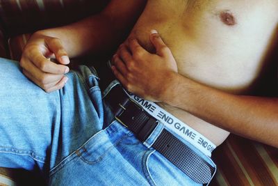 Midsection of shirtless man with hand on stomach lying at sofa