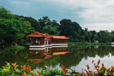 Built structure by lake against trees and plants against sky