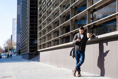 Man holding smartphone and coffee, standing next to office building