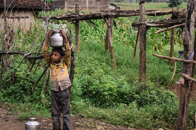 Boy carrying utensil on head standing by tree in forest