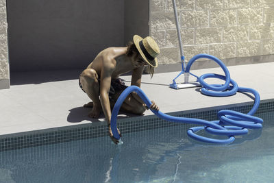 Young man wearing a hat crouched connecting the blue plastic pipe for the swimming pool cleaner