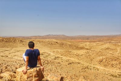 Rear view of young man sitting at desert against clear sky during sunny day