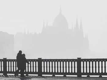 Silhouette couple walking on bridge against cathedral in foggy weather