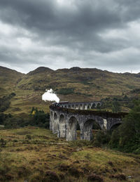 Arch bridge on field against cloudy sky