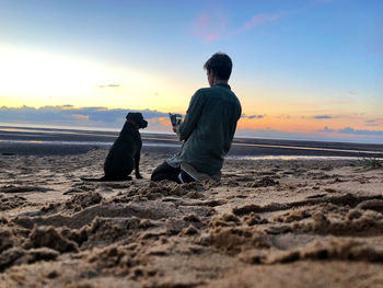 Woman photographing dog at beach during sunset