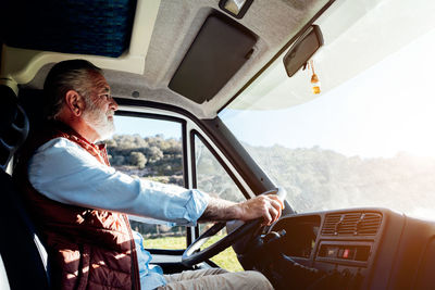 Side view of serious mature male traveler with beard driving automobile during road trip in countryside on sunny summer day